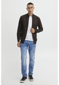Mørk melert zip-up cardigan med høyt krage, sammen med en hvit t-skjorte, lyseblå jeans og svarte joggesko. Løs passform og textured stoff.