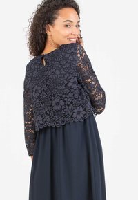 Marineblaues Kleid mit einer Spitzenüberlage, die florale Muster zeigt, langen Ärmeln und einem Schlüssellochdetail im Rücken. Der Rock ist glatt und fließend.