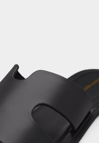 Κοντινή λήψη ενός μαύρου σανδαλιού χωρίς λουριά με ένα φαρδύ, λείο λουράκι που διαθέτει μια κομμένη εγκοπή σε απλό φόντο.