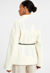 Vit, oversized jacka med hög krage och vida ärmar, bältad med ett svart band i midjan. Slät textur och minimalistisk design.