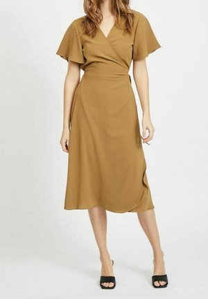 Kobieta w musztardowej sukience kopertowej z falbanianymi rękawami i czarnych sandałach na obcasie z odkrytymi palcami, stojąca na jasnym, jednolitym tle.