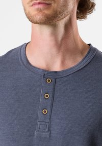 Maglietta a maniche lunghe di maglia lavorata a cialda blu con scollo rotondo e quattro bottoni marroni lungo la patta. Dettaglio del tessuto strutturato visibile.