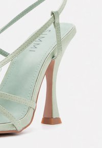 Πράσινο σουέτ πέδιλο με ψηλό τακούνι, λεπτά λουράκια που διασταυρώνονται πάνω από το πόδι και τετράγωνη μύτη.