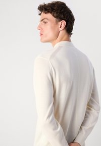 Maglia lunga a maniche con colletto alto di colore crema, texture liscia e dettagli delle cuciture sottili lungo la schiena e le maniche.