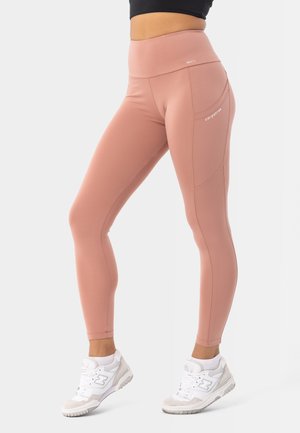 Světlé růžové legíny s vysokým pasem vyrobené z elastického materiálu, s bočními kapsami a bezšvovým designem, v kombinaci s bílými sportovními botami.