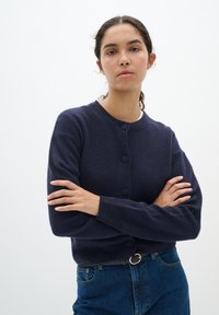 Marinblå stickad cardigan med rund halsringning, knäppning framtill, ribbade ärmslut och avslappnad passform, kombinerad med högmidjade jeans.