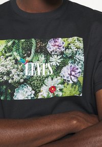 Černé bavlněné tričko s barevným grafickým motivem sukulentu, na kterém je vytištěno "LEVI'S" bílou barvou. Design zahrnuje různé textury a barvy rostlin.