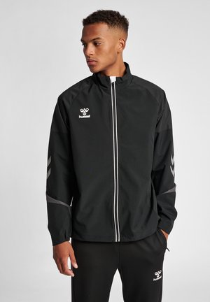 Junger Mann in schwarzer hummel Sportjacke mit weißem Reißverschluss und passenden schwarzen hummel Hosen, steht mit den Händen in den Taschen und schaut nach links.