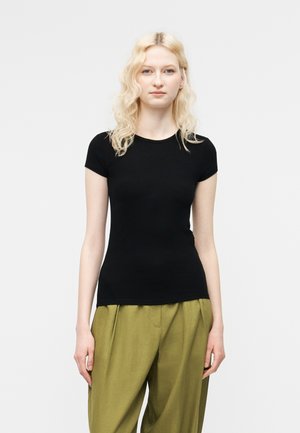 Ξανθιά γυναίκα που φοράει εφαρμοστό μαύρο κοντομάνικο τοπ και άνετο χακί παντελόνι, στέκεται μπροστά σε απλό φωτεινό φόντο.