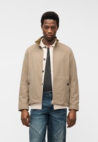 Άνδρας με στρωμένο μπεζ μπουφάν και πουκάμισο πάνω από μαύρο τοπ, φορώντας μπλε τζιν, στέκεται μπροστά σε απλό λευκό φόντο.