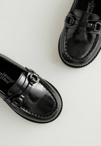 Mocasines de charol negro con un acabado suave, que presentan detalles metálicos y costuras elaboradas a lo largo de los bordes.