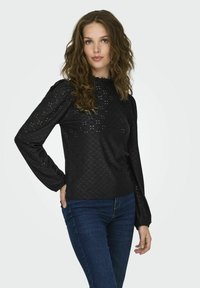 Black blouse with textured pattern and puffed sleeves. Features a high neckline and fitted cuffs. Paired with blue denim jeans.