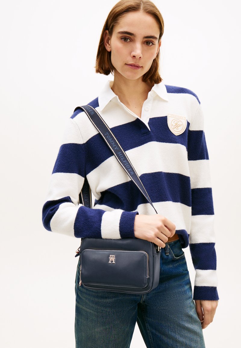 Tumesinine nahast õlakott ees oleva lukuga tasku, hõbedaste detailide ja rihmaga, millel on valge tekstiga kaubamärgi nimi.