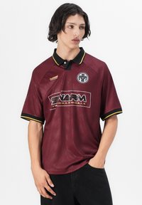 Jonge man die een kastanjebruine sportpolo draagt met een zwarte kraag, gele bies, logo's op de borst en een zwarte broek, poserend met één hand in de zak.