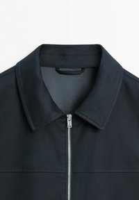 Giacca nera con cerniera, collo a punta, cuciture visibili e cursore della cerniera in metallo argentato al centro davanti.