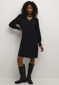 Robe noire mi-longue avec des manches longues froncées et un décolleté en V agrémenté d'un détail à nouer. Portée avec des bottes noires brillantes jusqu'au genou.