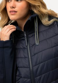 Giacca puffer navy con texture liscia, collo alto e zip a contrasto. Presenta dettagli verdi nei tiranti delle zip e nelle cuciture.