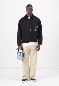 Svart denimjakke med frontknapper, sidelommer og logo-merke. Beige bukser og svarte joggesko. Holder en liten blå crossbody-veske.
