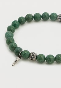 Βραχιόλι με πράσινες χάντρες, που αποτελείται από λείες, στρογγυλές πέτρες τύπου νεφρίτη με ασημένιες διακοσμητικές χάντρες που φέρουν τη λέξη "GUESS." Το κούμπωμα είναι λεπτομερές και μεταλλικό.