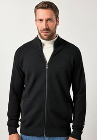 Giacca nera con zip e colletto alto, realizzata in tessuto morbido. Presenta polsini a coste e tasche laterali, abbinata a un dolcevita bianco sottostante.