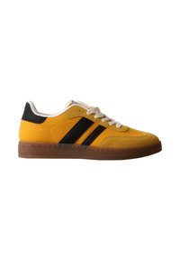 Sneaker jaune avec semelle marron, lacets blancs, patch noir au talon et deux bandes noires sur le côté, vu de l’extérieur.