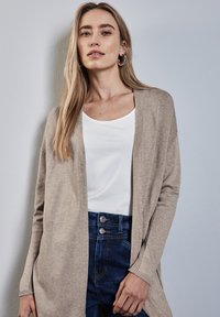 Beige Cardigan mit offenem Vorderteil, leichtes Strickmuster. Kombiniert mit einem weißen Oberteil und hochtaillierten, dunkelblauen Jeans mit dekorativen Knöpfen.
