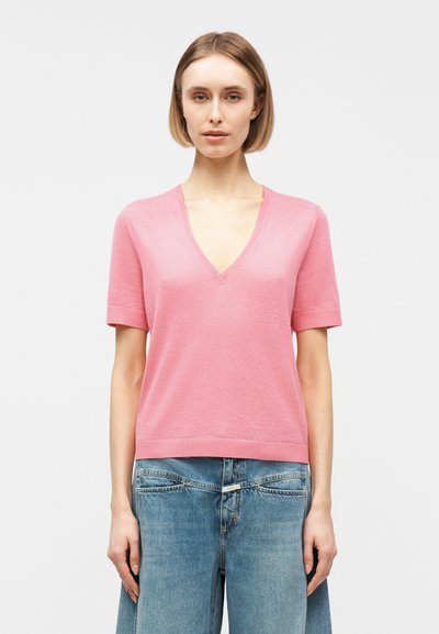 Femme portant un haut en maille rose à manches courtes et encolure en V, et un jean en denim bleu taille haute, debout devant un fond clair uni.