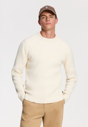 Cream ribbed knit sweater with a crew neck, worn over light brown corduroy pants. A small blue logo is visible on the sweater's hem.