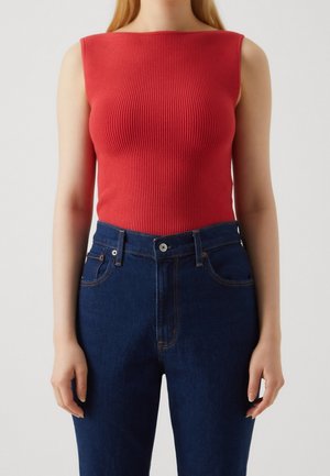 Vrouw die een mouwloze rode geribbelde top draagt, ingestopt in een hoge taille donkerblauwe spijkerbroek, tegen een effen achtergrond.