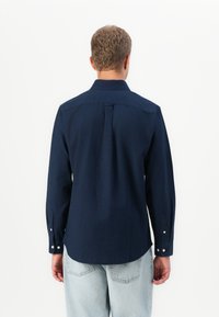 Hombre vistiendo una camisa de botones de manga larga azul marino y unos jeans de color azul claro, de pie con la espalda hacia la cámara frente a un fondo blanco liso.