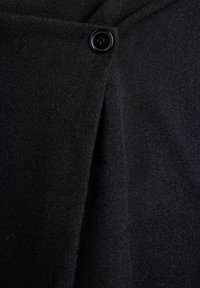Schwarzer Wollstoffmantel mit einem einzelnen glänzenden schwarzen Vierlochknopf, der oben in der Mitte befestigt ist.