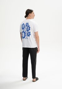 Witte katoenen t-shirt met een blauwe bloemenafbeelding en lachende gezichtjes op de achterkant, gecombineerd met zwarte broek en sandalen.