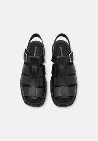 Sorte sandaler laget av glatt materiale, med et kryssdesign på stroppene, justerbar spenne og firkantet tåform med en strukturert fotseng.