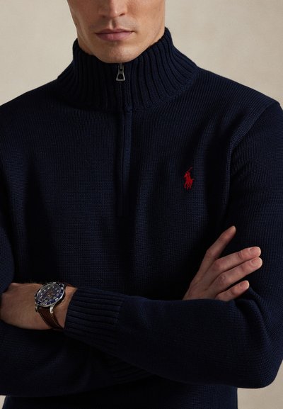 Mężczyzna z założonymi na siebie rękami, ubrany w ciemnogranatowy dzianinowy sweter na zamek z czerwonym logo konia oraz zegarek na rękę z brązowym skórzanym paskiem.