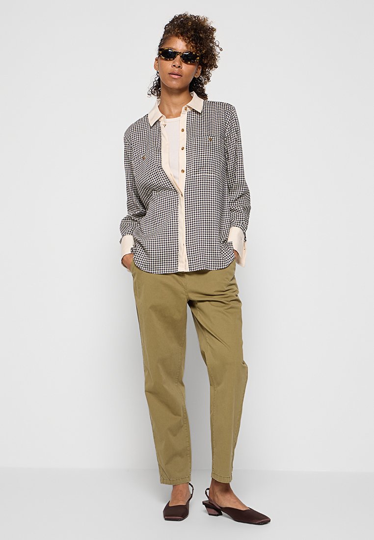 Frau trägt ein Hemd im Hahnentrittmuster, ein beiges Inneres, olivgrüne Hose, braune Slingback-Schuhe und dunkle Sonnenbrille, steht mit Händen in den Taschen.