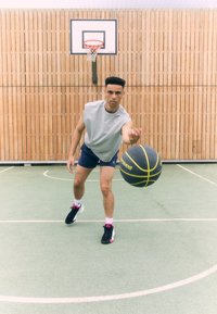 Balle de basket noire et jaune, débardeur gris sans manches, shorts bleus, chaussures noires avec des accents roses, sur un terrain avec un mur en bois et un panier visibles.