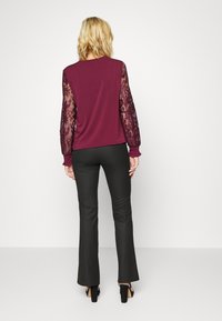 Bordeauxfarbener Sweatshirt mit langen Spitzenärmeln, kombiniert mit schwarzen Schlaghosen und Absatzschuhen. Glatte Textur mit gerafften Ärmelsäumen.