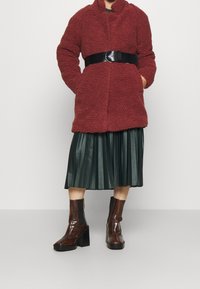 Texturerad röd fleecejacka med ett svart bälte, kombinerad med en plisserad grön fusk skinnkjol och bruna glansiga platformboots.