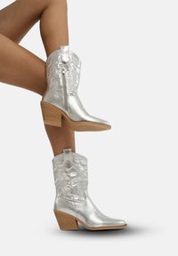 Srebrne metaliczne kowbojskie buty z ostrym noskiem, z detalami haftu na cholewie oraz z niskim obcasem. Zamek błyskawiczny po boku dla łatwego zakładania.