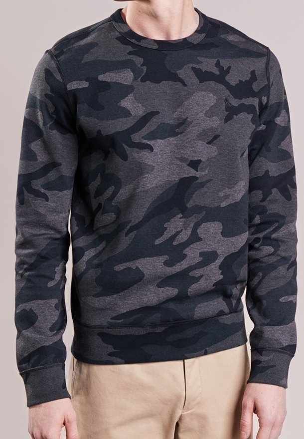 Homme portant un sweat-shirt à manches longues, à motifs camouflage gris foncé et noir, et un pantalon beige, debout devant un fond uni.