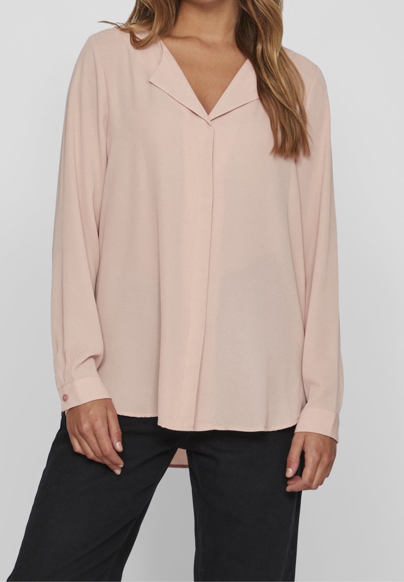 Femme portant un chemisier rose clair à manches longues avec un col en V et un pantalon noir, debout devant un fond gris clair uni.