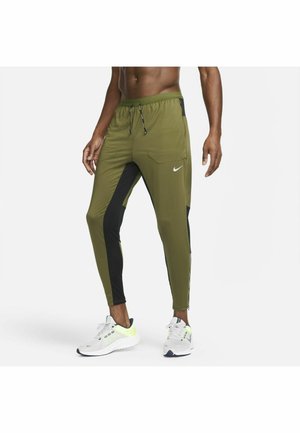 Homme portant un jogging vert et noir avec taille à cordon et des baskets blanches, debout devant un fond blanc uni.