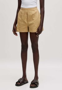 Khaki-Shorts mit strukturierter Oberfläche, elastischem Bund, Seitentaschen und lockerer Passform, kombiniert mit dunkelbraunen Sandalen.