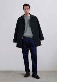 Abrigo de lana oscuro de doble botonadura sobre un suéter gris claro, combinado con pantalones azul oscuro y zapatos negros. Textura suave, ajuste entallado.