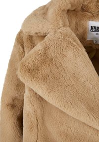 Kabát z umelej kožušiny v béžovej farbe, s výstrihom do V, mäkkou textúrou a prehnutými klopami. Na štítku je uvedené meno značky "Urban Classics" a veľkosť XS.