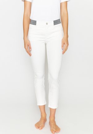 Vrouw die witte cropped broek draagt met een zwart-witte elastische tailleband, blootsvoets staand tegen een effen witte achtergrond.