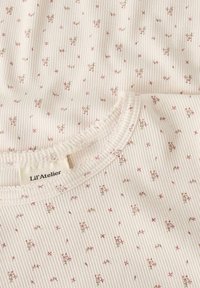 Zbliżenie beżowej żebrowanej tkaniny z małym różowym kwiecistym wzorem oraz falbaniastym wykończeniem dekoltu na garderobie Lil' Atelier.