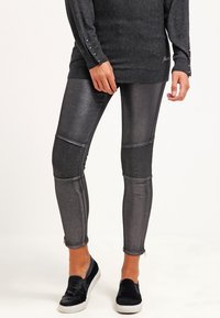 Leggings metálicos grises con paneles acanalados en las rodillas y dobladillos desgastados en los tobillos, combinados con una camiseta de manga larga oscura y zapatos negros tipo slip-on.