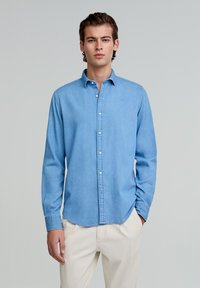Cămașă din denim albastru deschis, cu butoni și mâneci lungi, având un guler ascuțit și un logo subtil, asortată cu pantaloni de culoare crem.
