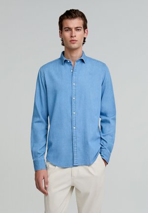 Lichtblauwe denim button-up shirt met lange mouwen, voorzien van een puntige kraag en een subtiel logo, gecombineerd met crèmekleurige broeken.